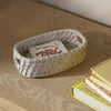 A small woven oval basket in a light natural beige color sits on a wooden tabletop, holding a playing card. A stack of books is partially visible on the right side.