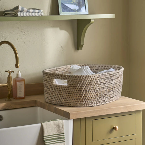 A round woven storage basket with cutout handles sits on a wooden countertop beside a sink in a styled bathroom or utility room. The basket contains folded white towels, and the surrounding environment includes a sage green cabinet, brass faucet, soap dispenser, and shelf.