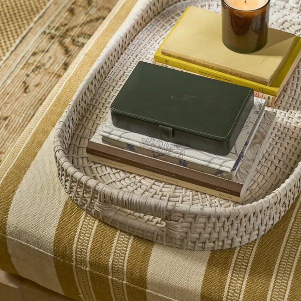 A woven oval tray holding stacked books and a dark green leather case, styled on a striped beige and olive textile surface with a candle nearby.