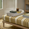 A woven oval tray holding stacked books and a small amber candle sits on a striped upholstered bench in a softly lit room.