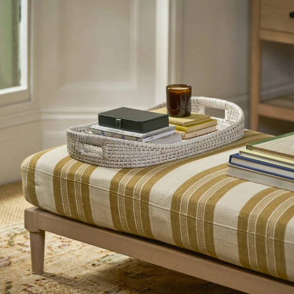 A woven oval tray holding stacked books and a small amber candle sits on a striped upholstered bench in a softly lit room.