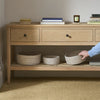 A light natural wood console table with drawers and an open shelf, styled with books and woven storage baskets. A person's arm and hand are visible holding one of the baskets on the lower shelf in a softly lit room.