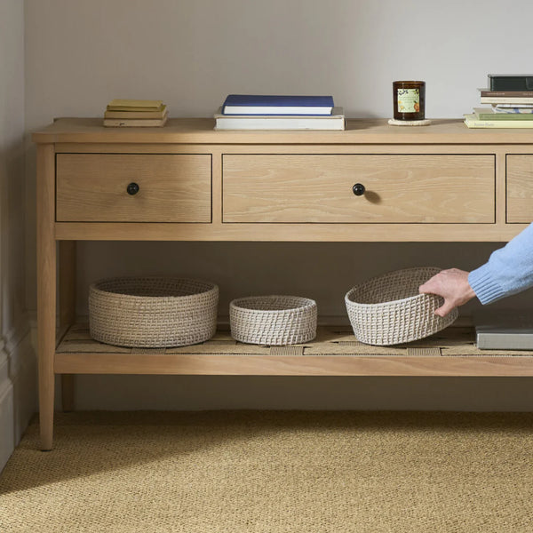 A light natural wood console table with drawers and an open shelf, styled with books and woven storage baskets. A person's arm and hand are visible holding one of the baskets on the lower shelf in a softly lit room.