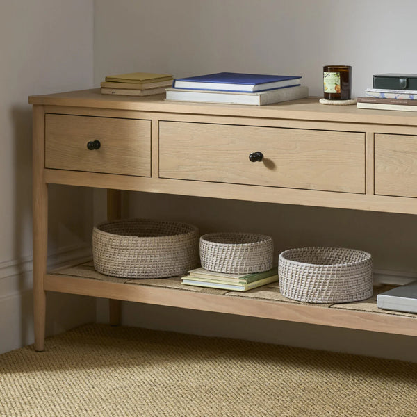 A light natural wood console table with three drawers and a lower shelf, styled with books, baskets, and a candle in a neutral room corner.