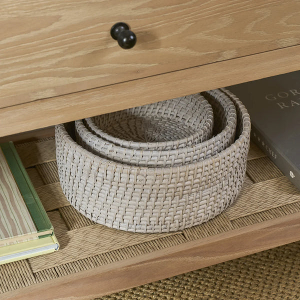 A light beige woven basket tray nested on a wooden shelf under a cabinet, with books partially visible on either side.