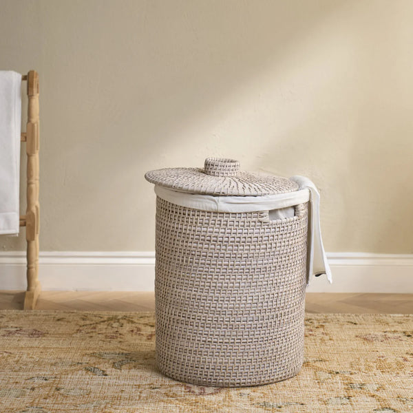 A woven cylindrical laundry hamper with a matching lid, shown in a styled room setting with a beige wall, patterned rug, and a wooden towel rack on the left.