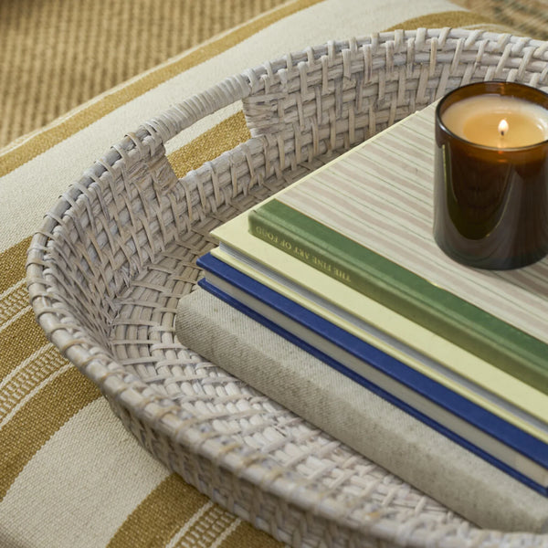A close-up of a woven rounded square tray holding stacked books and a lit amber glass candle, placed on a striped beige and tan textile surface.