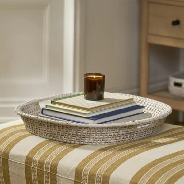 A rounded square woven white tray holding stacked books and a small amber glass candle, placed on a striped upholstered bench in a softly lit indoor room.