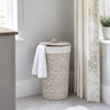 A woven cylindrical laundry hamper with a matching lid, shown in a softly styled room corner beside a window and curtain. A white towel is draped over the side of the hamper.