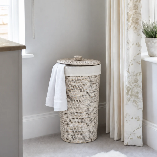 A woven cylindrical laundry hamper with a matching lid, shown in a softly styled room corner beside a window and curtain. A white towel is draped over the side of the hamper.