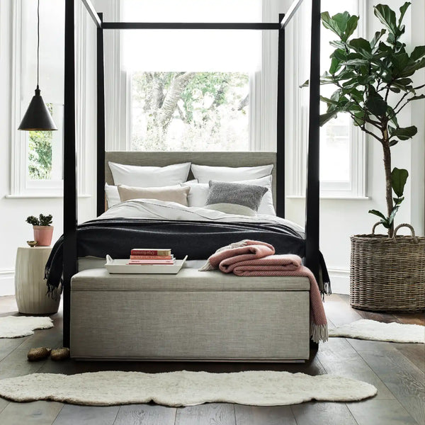 A modern bedroom scene featuring a large sheepskin rug, four-poster canopy bed with a light gray upholstered bench at the foot, styled with neutral bedding, a pink throw blanket, and a large potted fiddle-leaf fig plant in a woven basket beside the window.
