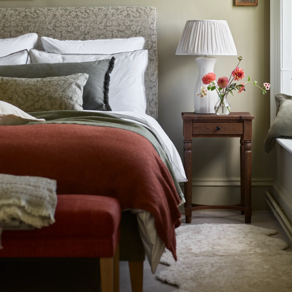A styled bedroom scene featuring a sheepskin rug, bed with white and gray patterned bedding, a rust-red textured throw blanket, a matching upholstered bench in the foreground, and a dark wood nightstand holding a white pleated table lamp with flowers in a glass vase.