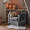 A sheepskin rug, dusty blue-gray upholstered armchair with matching throw pillows and a draped throw blanket, styled in a cozy attic reading nook with a floor lamp, bookshelves, and a slanted ceiling.