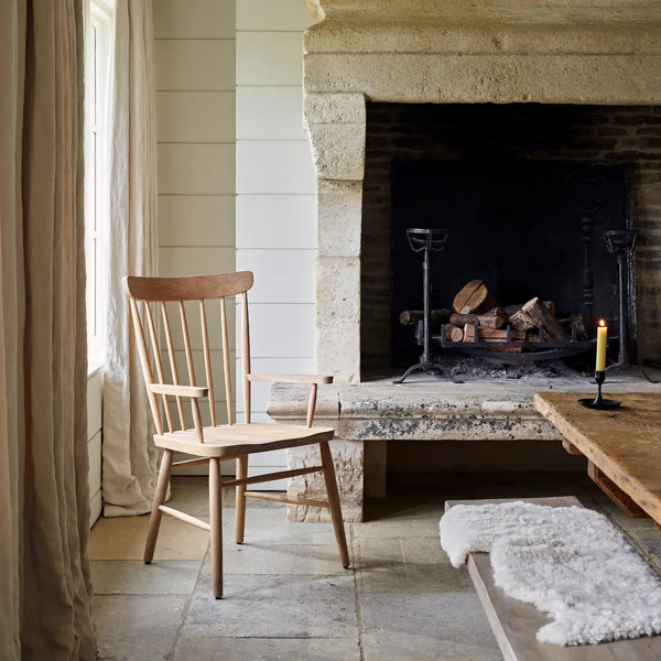 A light natural wood spindle-back chair sits in a rustic room beside a stone fireplace with a candle on a table and a white sheepskin rug on the bench.