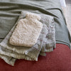 A folded cream sherpa hot water bottle cover resting on a textured gray throw blanket with fringe, which is laid over a rust-red upholstered surface.