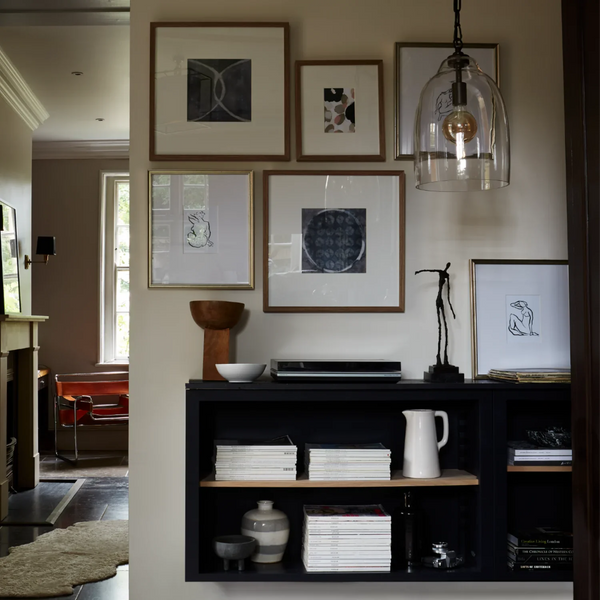 A styled interior entryway featuring a gallery wall with five framed art prints in light wood frames, a black floating shelf with books and ceramic vessels, and a clear glass bell pendant light hanging above.