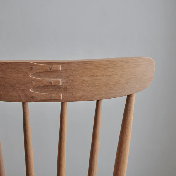 Close-up of a light natural wood chair backrest showing the carved joinery and spindle legs against a plain gray background.