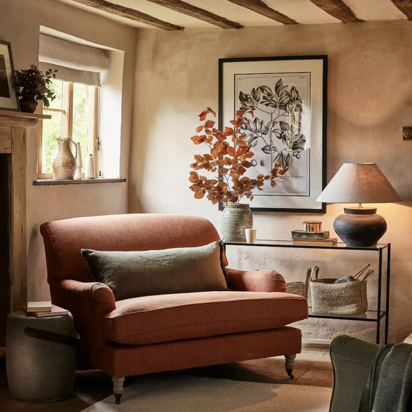 Framed botanical art on a plaster wall in a warm, rustic living room scene featuring a terracotta-colored upholstered armchair with a sage green throw pillow, set next to a black metal side table with a lamp.