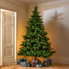 A tall artificial Christmas tree stands in the corner of a room with beige walls and carpet, with wrapped gift boxes placed at its base. A doorway is visible on the left, and warm light casts a tree-like shadow on the wall.