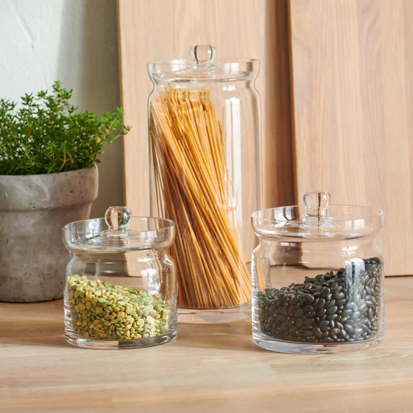 Three clear glass canisters with glass lids and small round handles, arranged on a wooden surface. The tallest canister holds spaghetti sticks, while the shorter canisters contain green lentils and dark beans.