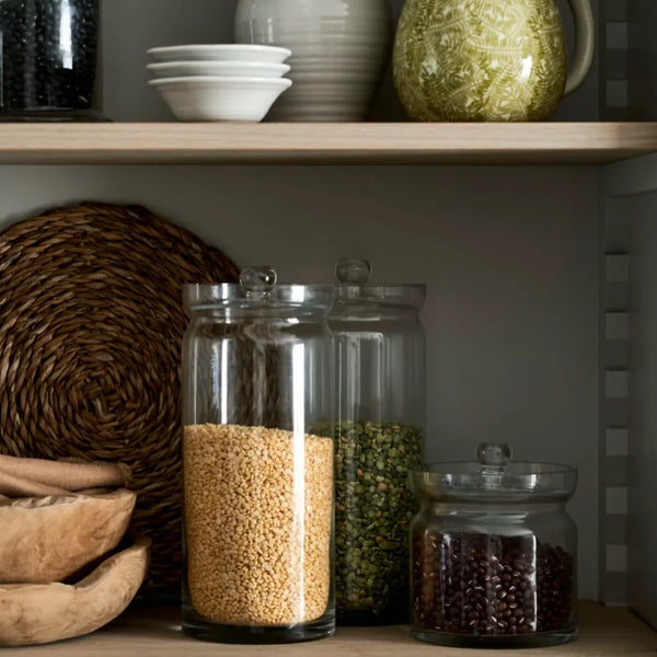 Three clear glass canisters with lids, filled with grains, green beans, and dark red beans respectively, arranged on a wooden shelf next to a woven placemat.