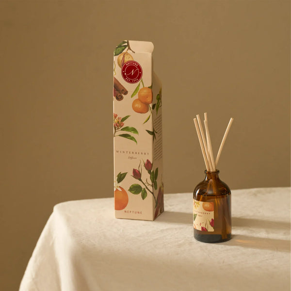 A reed diffuser bottle with amber glass and natural reeds sits beside its illustrated product box on a light fabric surface. The packaging and bottle label show winterberry, citrus, cinnamon, and floral illustrations, with the brand name Nature's Own visible.