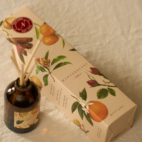 A reed diffuser bottle with amber glass and natural reeds sits beside its illustrated product box on a light fabric surface. The packaging and bottle label show winterberry, citrus, cinnamon, and floral illustrations, with the brand name Nature's Own visible.