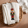 A small embroidered nutcracker ornament hangs from a black cord over a stack of white napkins on a woven round placemat, with a wooden table and white fabric in the background.