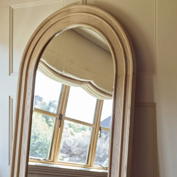 An arched full-length wooden floor mirror with a natural oak finish, leaning against a cream paneled wall with a window visible in its reflection.