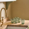 Six green-tinted glass tumblers with a textured, crackled finish arranged on a wooden kitchen counter next to a brass faucet.