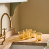 Five amber glass tumblers with a textured, crackled finish arranged on a wooden kitchen countertop next to a brass faucet.