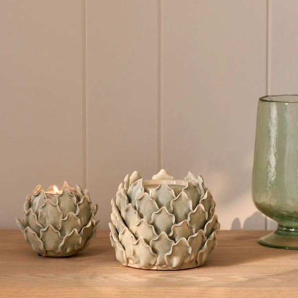 Two ceramic artichoke-shaped candle holders on a wooden surface against a beige paneled wall, with one holder lit and a green glass vessel partially visible on the right.