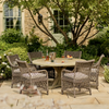 An outdoor patio dining set featuring a round woven wicker table and six matching armchairs with light cushions, arranged on a stone patio in front of a brick house with greenery.