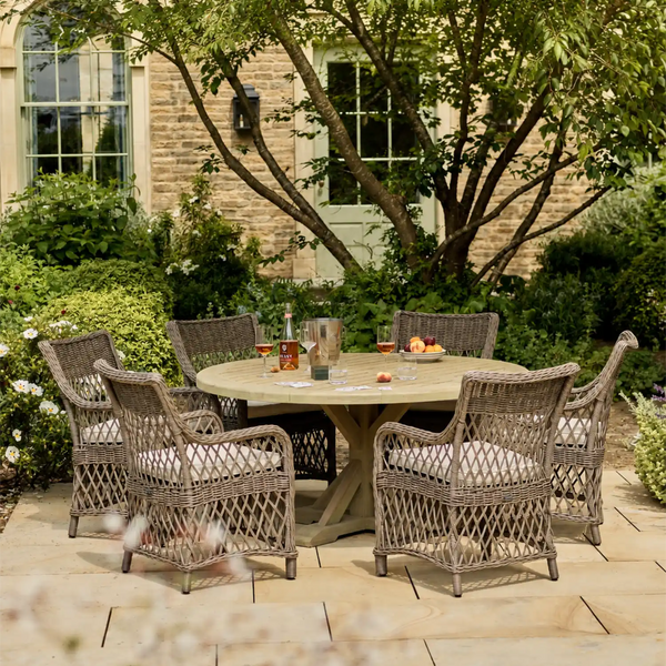 An outdoor patio dining set featuring a round woven wicker table and six matching armchairs with light cushions, arranged on a stone patio in front of a brick house with greenery.
