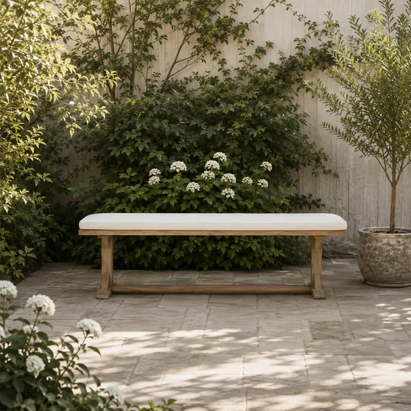 A rectangular outdoor bench with a light beige cushion and natural wood frame, set on a stone patio in a garden with green foliage and white flowers.