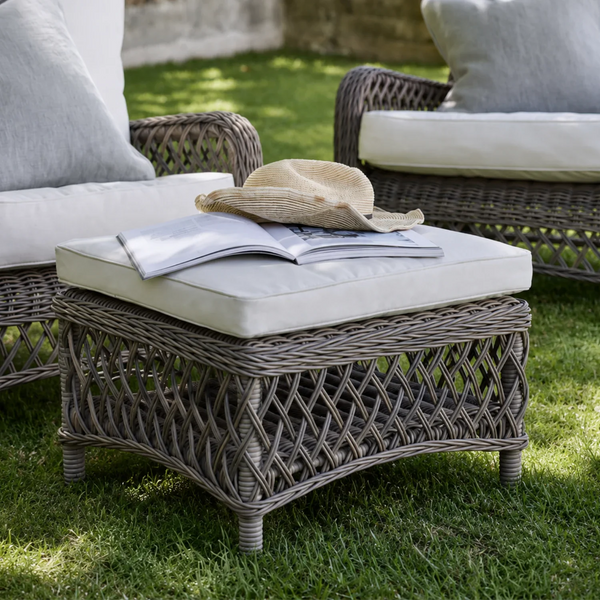 A square outdoor wicker ottoman with a cream cushion sits on a green lawn, styled with an open book and straw hat. Matching patio chairs are partially visible in the background.