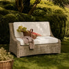 Outdoor wicker patio loveseat with white cushions in a garden setting, styled with a basket of vegetables and a folded blanket.