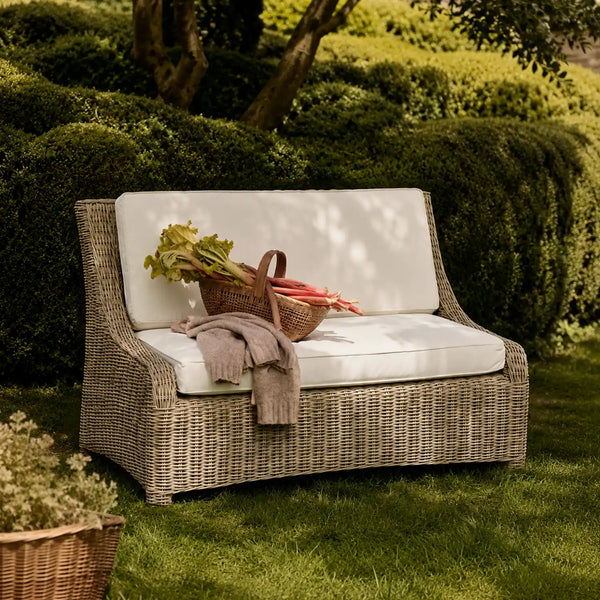 Outdoor wicker patio loveseat with white cushions in a garden setting, styled with a basket of vegetables and a folded blanket.