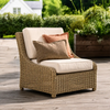 A wicker outdoor armchair with cream cushions and two decorative pillows, set on a wooden deck with garden greenery in the background.