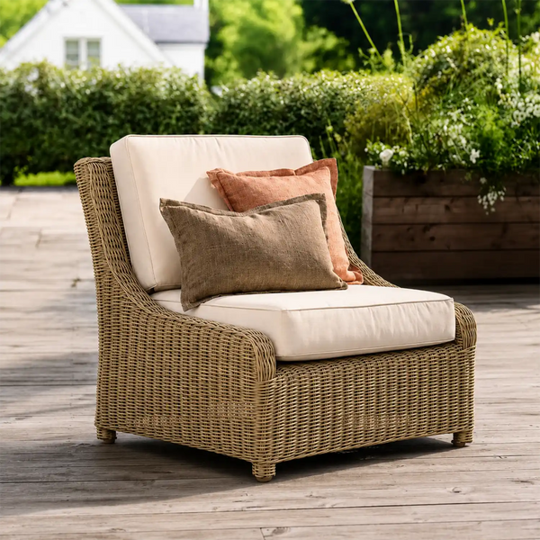 A wicker outdoor armchair with cream cushions and two decorative pillows, set on a wooden deck with garden greenery in the background.