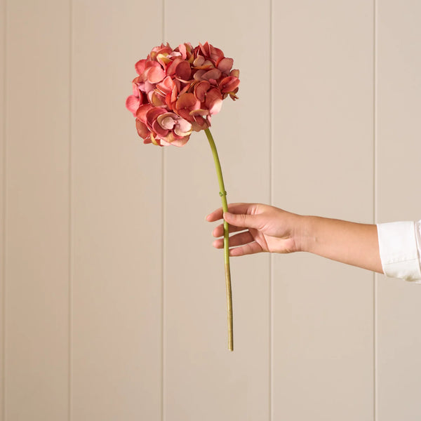 A person's hand holding a single long-stemmed artificial hydrangea with dusty pink and coral-orange blooms against a beige paneled wall.
