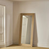 A tall floor mirror with a light natural wood frame leaning against a cream-colored wall in a room with carpeted floor and white trim.