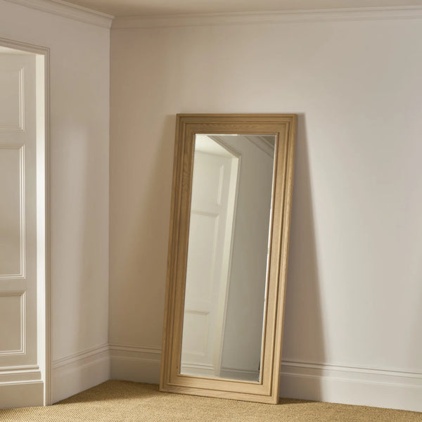 A tall floor mirror with a light natural wood frame leaning against a cream-colored wall in a room with carpeted floor and white trim.