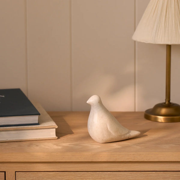 A small sculptural bird figurine in a warm off-white stone or ceramic finish, displayed on a light wood sideboard next to a stack of books and a table lamp.