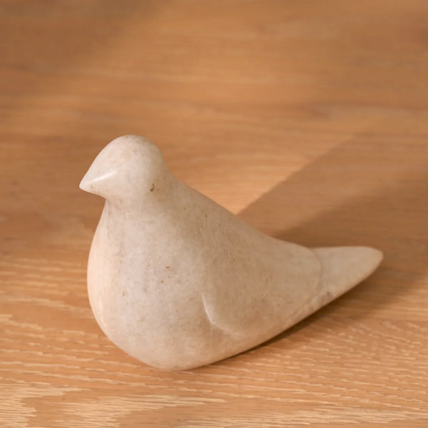 A small sculpted bird figurine made of light beige stone, resting on a warm-toned wooden surface.