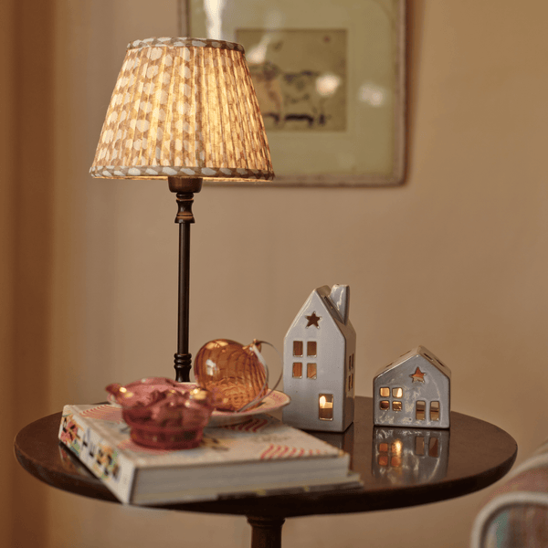 A lit table lamp with a beige patterned shade and dark stem stands on a round wooden side table beside two small white house-shaped ceramic holders with warm glowing windows, along with books and a pink bowl in a softly styled interior setting.