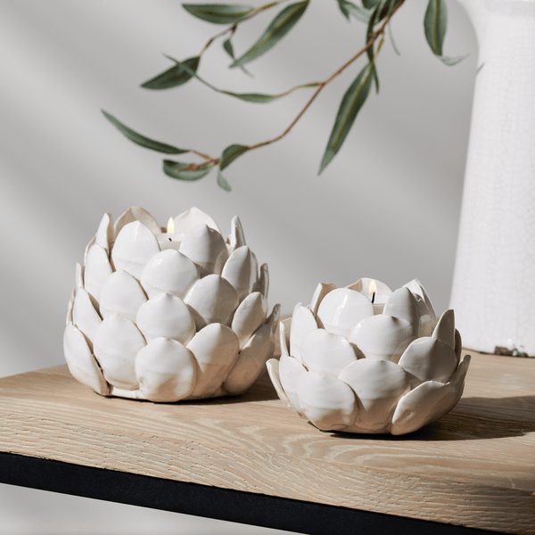 Two white ceramic artichoke-shaped candle holders with a lit tea light inside, displayed on a light wood surface with a neutral background and soft shadows.