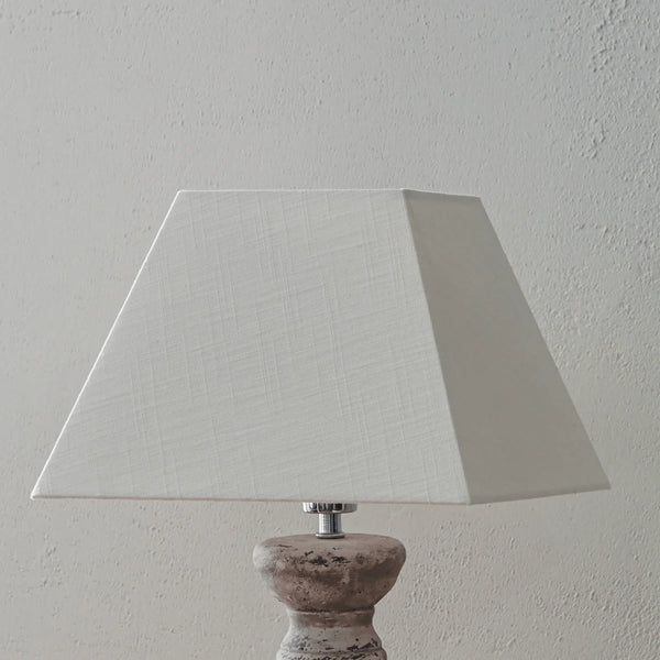 A table lamp with a large white textured fabric drum shade and a distressed gray-brown wooden base, shown against a textured off-white wall.