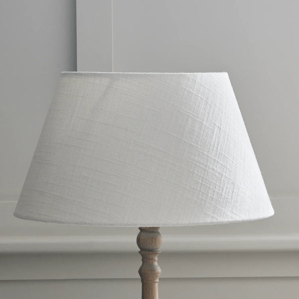 A white textured fabric drum lampshade mounted on a light-colored wooden lamp base, set against a neutral gray wall.