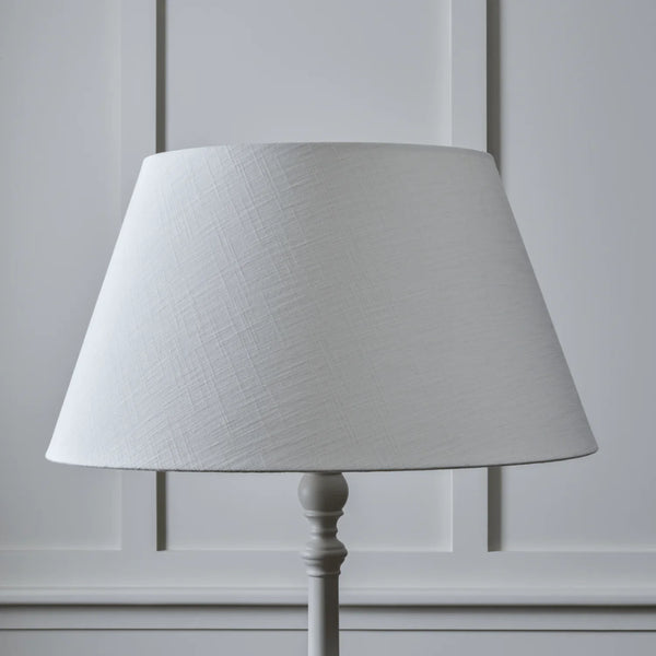 A white fabric drum lampshade with a subtle woven texture, mounted on a white painted metal floor lamp stand against a white paneled wall.
